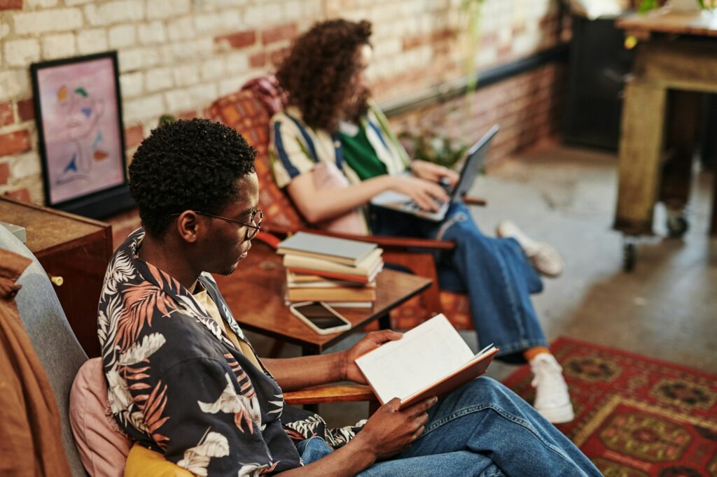 African American guy reading book while sitting in armchair in living room